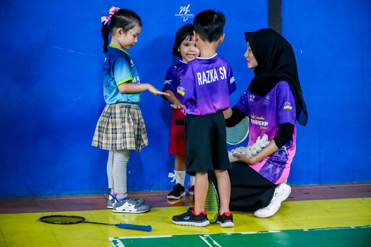 Asisten pelatih berjongkok berbincang dengan tiga anak kecil di pinggir lapangan